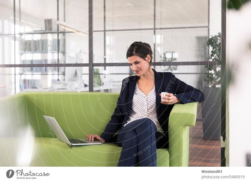 Geschäftsfrau mit Laptop, die auf einer Couch sitzt und im Büro Kaffee trinkt Europäer Kaukasier Europäisch kaukasisch Dreiviertelansicht dunkelhaarig