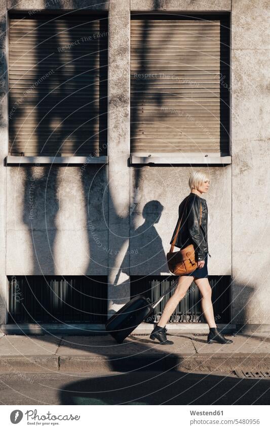 Blonde Frau mit Gepäck geht auf dem Bürgersteig Europäer Kaukasier Europäisch kaukasisch unterwegs auf Achse in Bewegung Seitenansicht von der Seite gehen