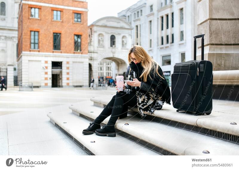 UK, London, junge Frau mit Gepäck, die ihr Handy auf der Treppe überprüft Papierbecher Pappbecher Becher SMS Textnachricht Städtereise City Trip Kurztripp