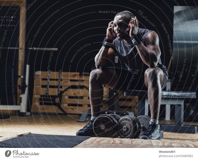 Athlet sitzt im Fitnessstudio, trägt Kopfhörer, konzentriert sich Gewicht Gewichte Muskeln muskulös athletisch Fitnessclubs Fitnessstudios Turnhalle Sportler