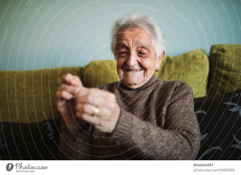 Porträt einer lächelnden älteren Frau, die auf der Couch sitzt und den Faden durch das Knopfloch einer Nähnadel führt Seniorin Seniorinnen alt Portrait Porträts