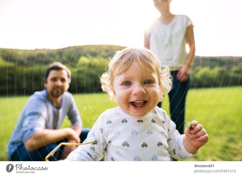 Süßer kleiner Junge auf der Wiese mit den Eltern im Hintergrund Baby Babies Babys Säuglinge Kind Kinder Spaß Spass Späße spassig Spässe spaßig lachen Portrait