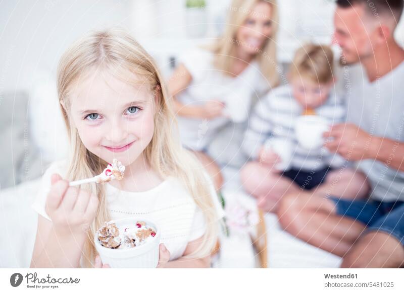 Porträt eines lächelnden Mädchens beim Essen aus der Müslischale mit Familie im Hintergrund Portrait Porträts Portraits weiblich essen essend Kind Kinder Kids