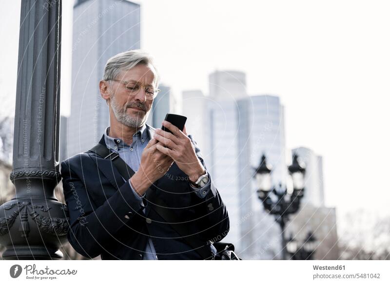 Grauhaariger Geschäftsmann betrachtet Smartphone neben Straßenlampe stehend Businessmann Businessmänner Geschäftsmänner Handy Mobiltelefon Handies Handys