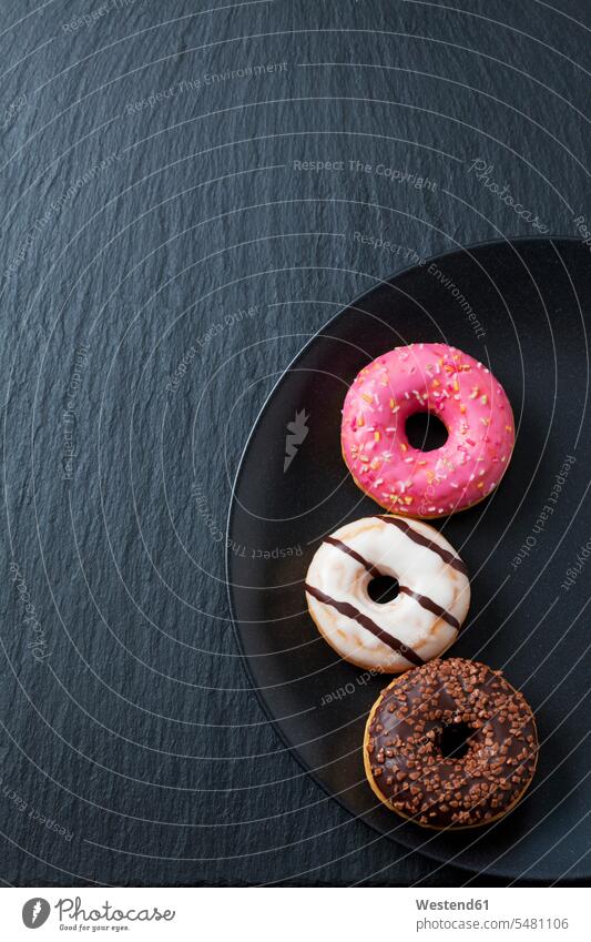 Drei Doughnuts mit unterschiedlicher Glasur auf Schwarzblech und Schiefer Gruppierung Anordnung Ordnung gruppiert gruppieren süß Süßes rund Rundungen Donut