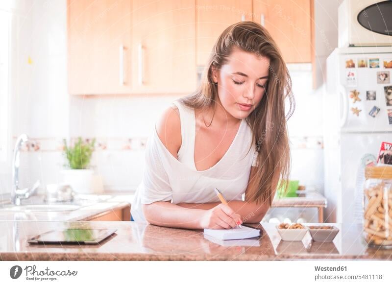 Junge Frau schreibt auf Notizblock in Küche Notizbloecke Notizblöcke schreiben aufschreiben notieren schreibend Schrift weiblich Frauen Erwachsener erwachsen