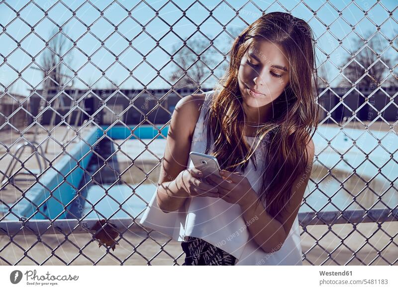 Attraktive junge Frau an einem Zaun, die auf ihr Handy schaut Mobiltelefon Handies Handys Mobiltelefone weiblich Frauen Telefon telefonieren Kommunikation