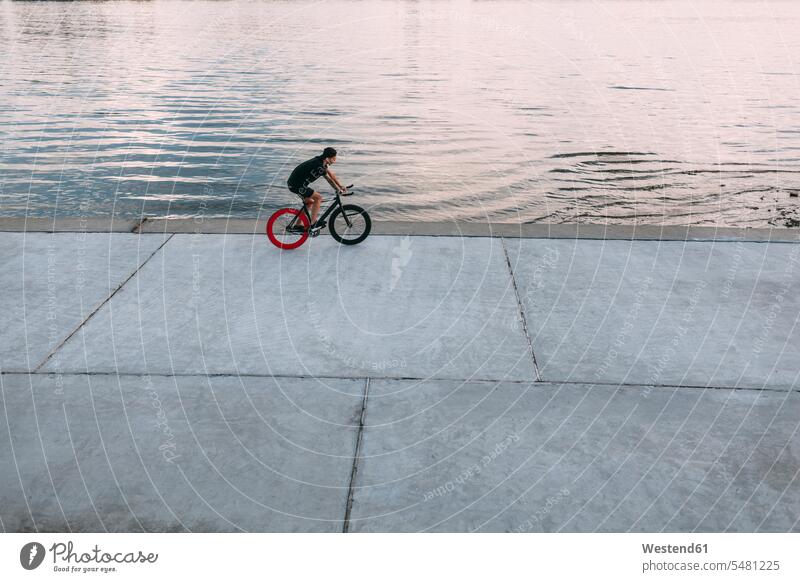 Junger Mann fährt Fixie-Rad am Wasser Fluss Fluesse Fluß Flüsse fahren fahrend fahrender fahrendes Männer männlich Fahrrad Bikes Fahrräder Räder Gewässer