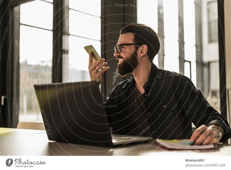 Junger Gelegenheits-Geschäftsmann sitzt im Büro und hinterlässt mit seinem Smartphone eine Voice-Mail Laptop Notebook Laptops Notebooks sprechen reden