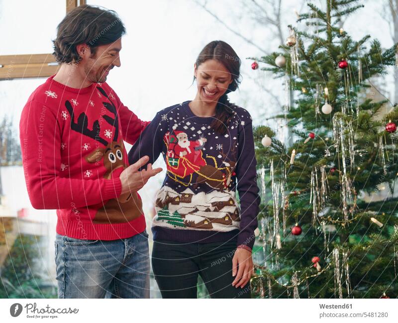 Paar, das vor dem Weihnachtsbaum steht und Weihnachtspullover trägt Europäer Kaukasier Europäisch kaukasisch Strickpullover Überraschung überraschen stehen
