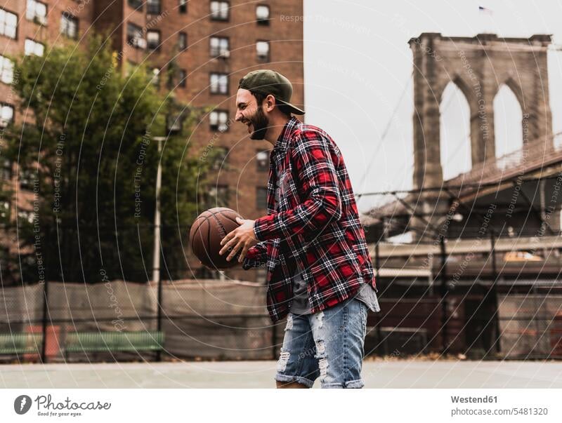 USA, New York, lachender junger Mann mit Basketball auf einem Aussenplatz trainieren spielen Basketbaelle Basketbälle Männer männlich Sport positiv Emotion