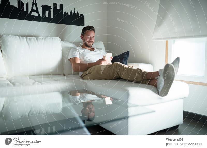 Lächelnder junger Mann entspannt zu Hause mit seinem Smartphone Zuhause daheim lächeln Sofa Couches Liege Sofas sitzen sitzend sitzt Handy Mobiltelefon Handies