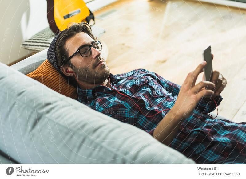 Junger Mann liegt auf der Couch und hört Musik mit Kopfhörern und Smartphone Hemd Hemden aufstützen aufgestuetzt aufgestützt Rückenlage auf dem Rücken