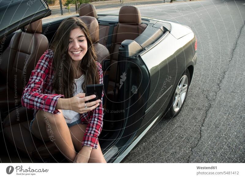 Lächelnde junge Frau sitzt im Cabrio und schaut auf Smartphone Kabriolett Kabrios Cabriolet Cabriolets Kabrioletts Cabrios weiblich Frauen lächeln sitzen