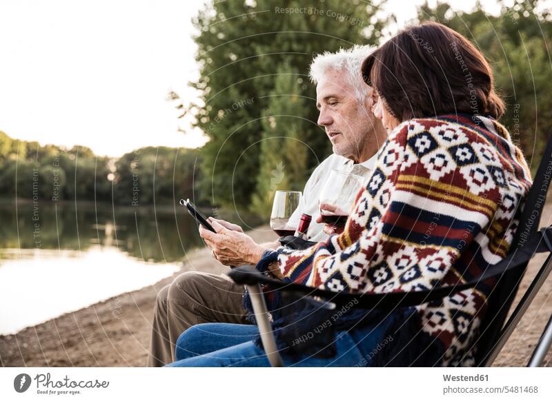 Älteres Ehepaar mit Wein sitzend an einem See mit Tablette Seen entspannt entspanntheit relaxt Paar Pärchen Paare Partnerschaft Tablet Computer Tablet-PC