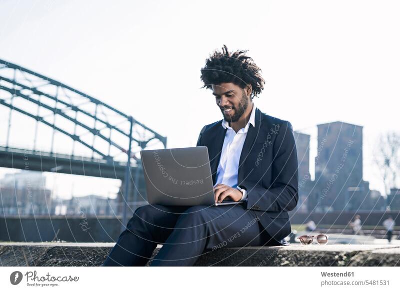 Lächelnder Geschäftsmann sitzt mit Laptop am Flussufer Notebook Laptops Notebooks Mann Männer männlich Businessmann Businessmänner Geschäftsmänner lächeln