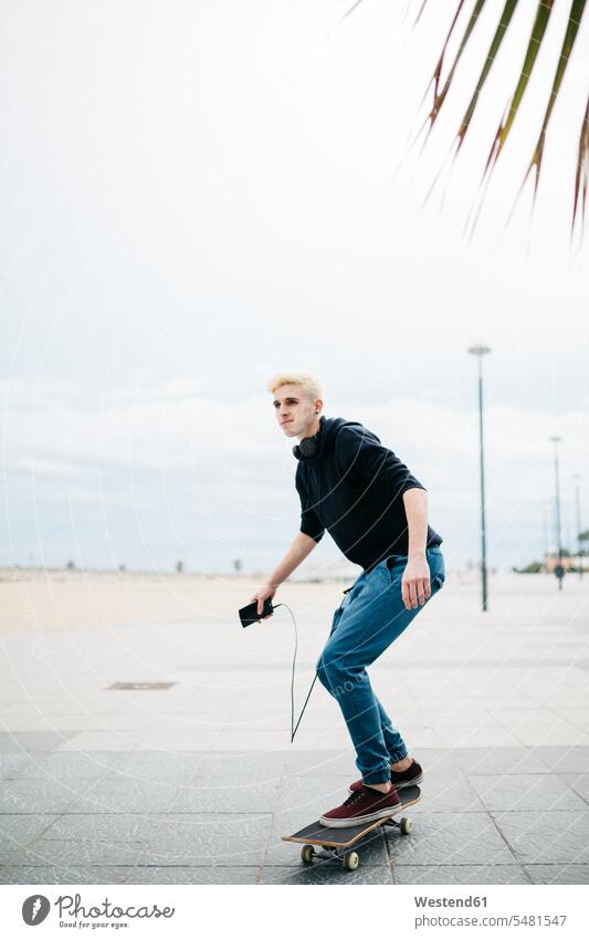 Spanien, Torredembarra, junger Mann mit Smartphone auf seinem Skateboard stehend Europäer Kaukasier Europäisch kaukasisch unterwegs auf Achse in Bewegung Agil