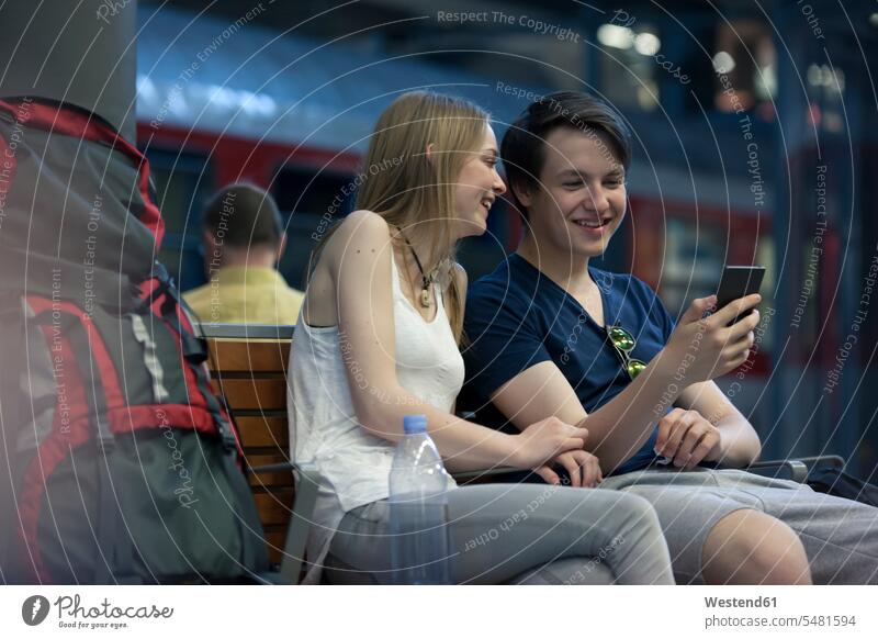 Junges Paar mit Rucksäcken sitzt mit Smartphone am Bahnhof Pärchen Paare Partnerschaft Reisende Reisender Rucksacktourist Backpacker iPhone Smartphones