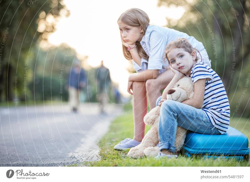 Zwei Mädchen sitzen auf einem Koffer am Straßenrand warten sitzend sitzt Schwester Schwestern Straßenränder Strassenrand Strassenraender Geschwister Familie