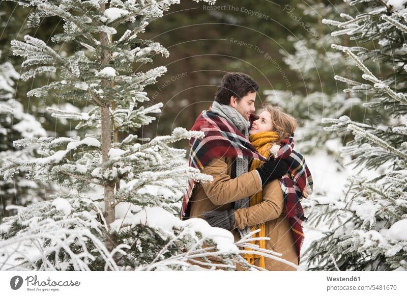 Glückliches junges Paar in eine Decke gehüllt im Winterwald stehend Pärchen Paare Partnerschaft Schnee Mensch Menschen Leute People Personen Wetter ansehen