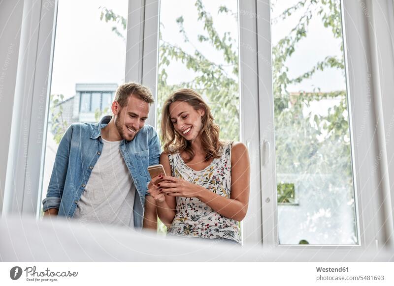 Lächelndes Paar vor dem Fenster, das auf ein Smartphone schaut Pärchen Paare Partnerschaft iPhone Smartphones lächeln Mensch Menschen Leute People Personen