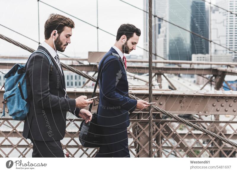 USA, New York City, zwei Geschäftsleute mit Mobiltelefonen und Ohrstöpseln auf der Brooklyn Bridge Geschäftsmann Businessmann Businessmänner Geschäftsmänner