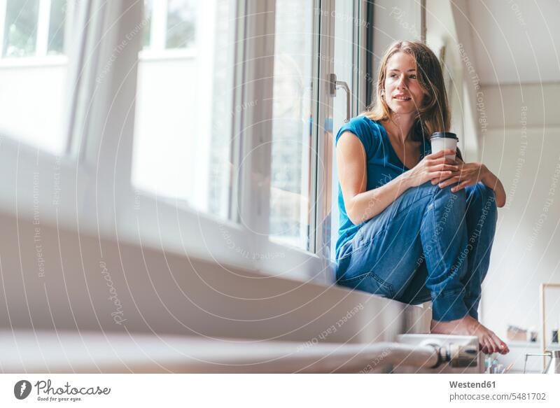 Lächelnde junge Frau mit Kaffee zum Mitnehmen am Fenster sitzend weiblich Frauen lächeln entspannt entspanntheit relaxt Getränk Getraenk Getränke Getraenke