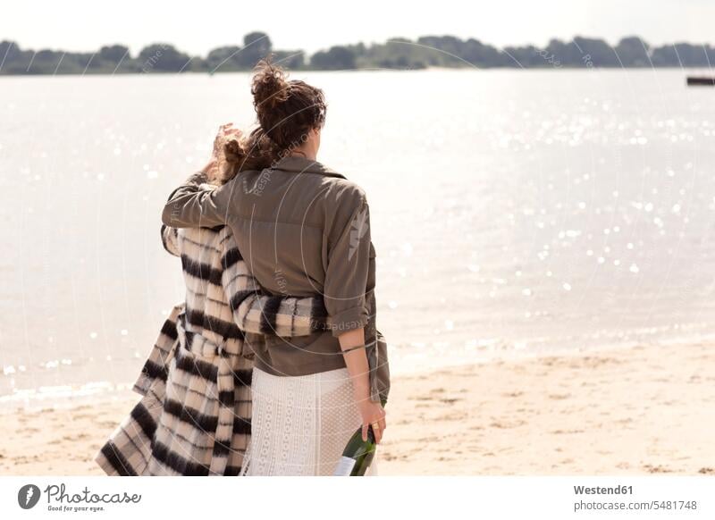 Rückenansicht von zwei Freunden, die Arm in Arm mit einer Flasche Sekt am Strand spazieren gehen Freundinnen Beach Straende Strände Beaches Freundschaft