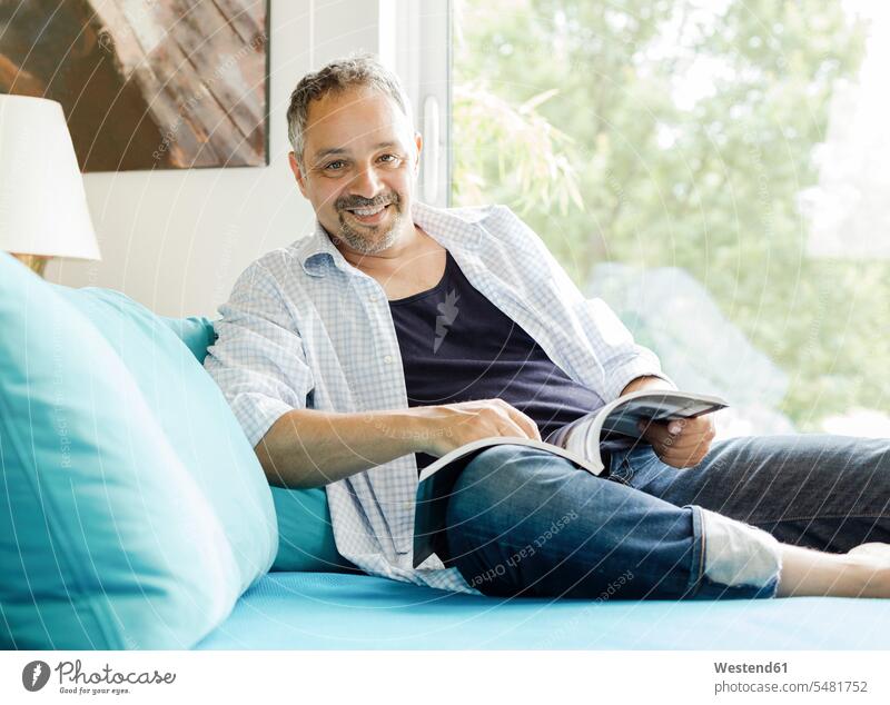 Porträt eines lächelnden Mannes auf der Couch sitzend mit Magazin Männer männlich Sofa Couches Liege Sofas Erwachsener erwachsen Mensch Menschen Leute People