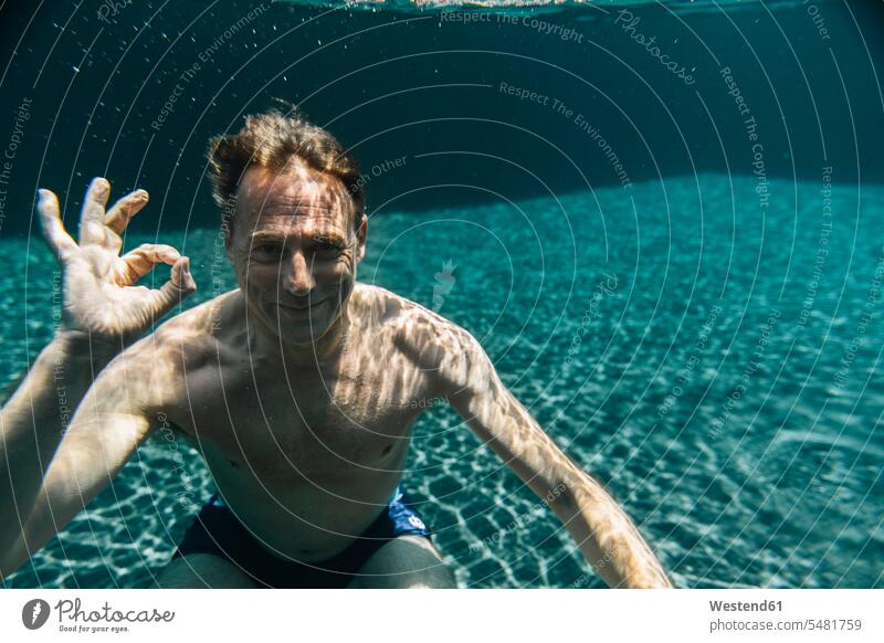 Porträt eines lächelnden Mannes unter Wasser in einem Schwimmbad, der ein Ok-Zeichen macht Swimmingpool Swimmingpools Schwimmbecken Swimming Pool Swimming Pools