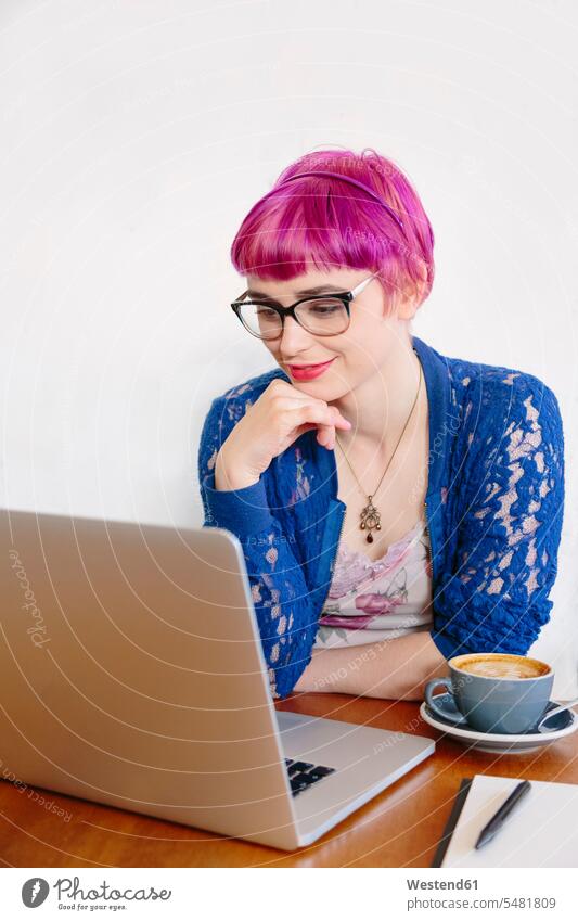 Porträt einer lächelnden jungen Frau mit gefärbten Haaren, die in einem Café sitzt und auf einen Laptop schaut Notebook Laptops Notebooks weiblich Frauen