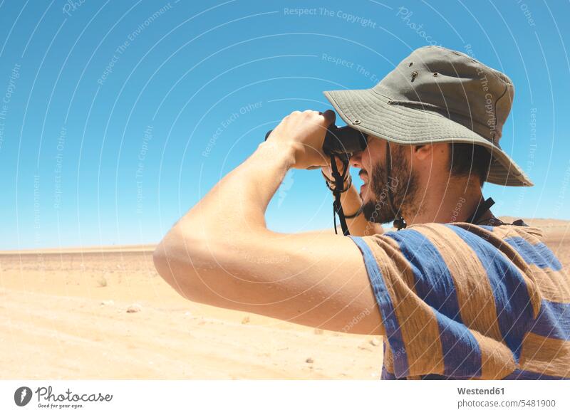 Namibia, Namib-Wüste, Mann mit Hut, der mit dem Fernglas wegschaut Europäer Kaukasier Europäisch kaukasisch Naturtourismus Käppi Mütze Kappe Kaeppis Mützen