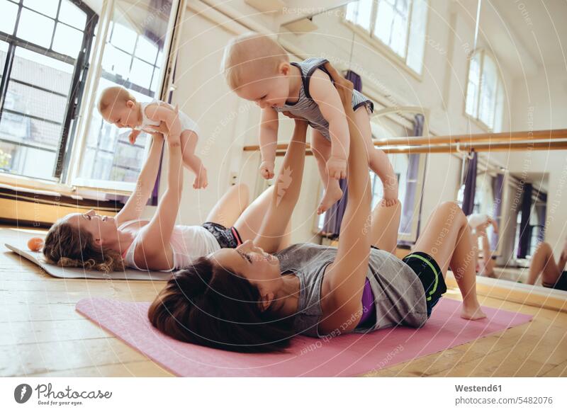 Zwei Mütter, die auf Yogamatten trainieren, während sie ihre Babys hochhalten Mutter Mami Mutti Mama lächeln heben Spaß Spass Späße spassig Spässe spaßig Babies