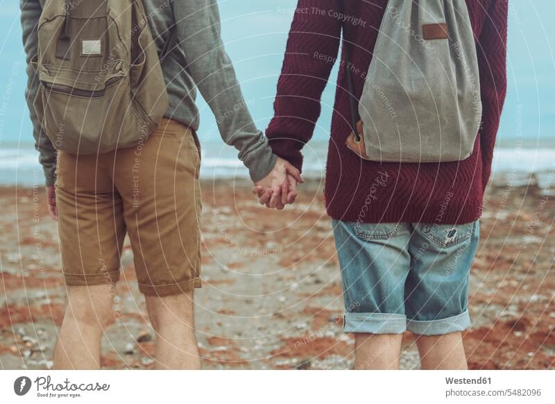 Rückenansicht eines jungen schwulen Paares, das sich am Strand an den Händen hält Pärchen Partnerschaft Beach Straende Strände Beaches Mensch Menschen Leute