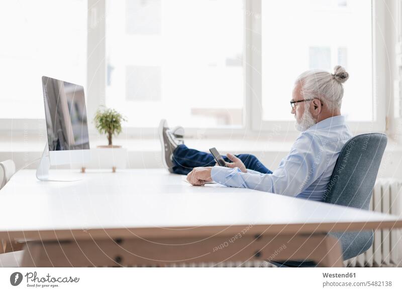 Reifer Mann mit Bart benutzt Mobiltelefon am Schreibtisch Männer männlich Pause entspannt entspanntheit relaxt Erwachsener erwachsen Mensch Menschen Leute
