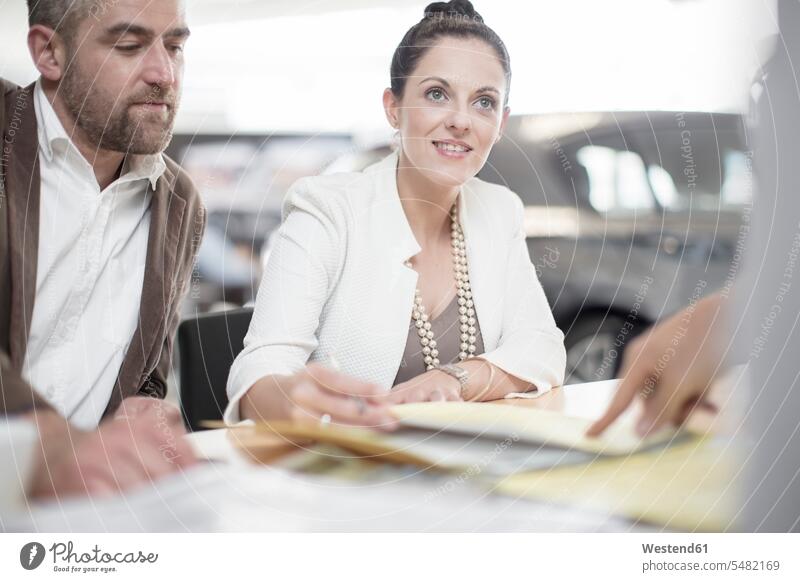 Autohändler und Ehepaar sitzen am Schreibtisch mit Vertragsformularen im Ausstellungsraum Autohaendler Autohaus Autohäuser Autohaeuser Autohandlung
