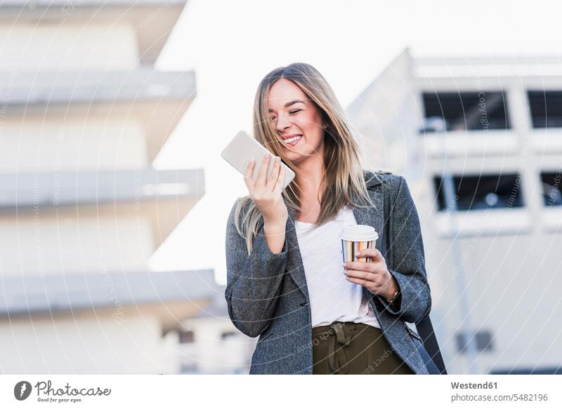 Glückliche junge Frau mit Kaffee zum Mitnehmen und Handy in der Stadt staedtisch städtisch glücklich glücklich sein glücklichsein Mobiltelefon Handies Handys