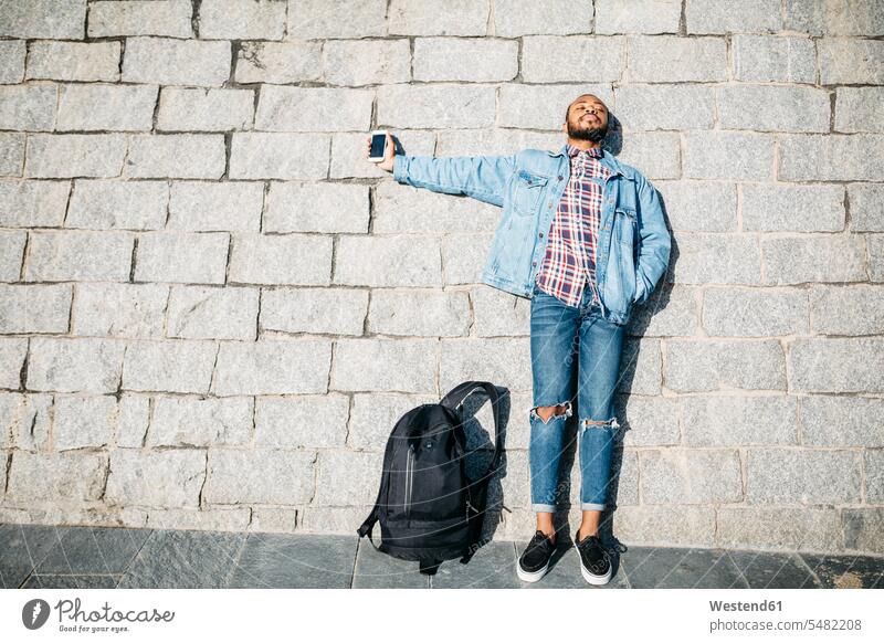 Junger Mann steht mit einem Handy vor einer Wand Smartphone iPhone Smartphones stehen stehend Männer männlich Mobiltelefon Handies Handys Mobiltelefone