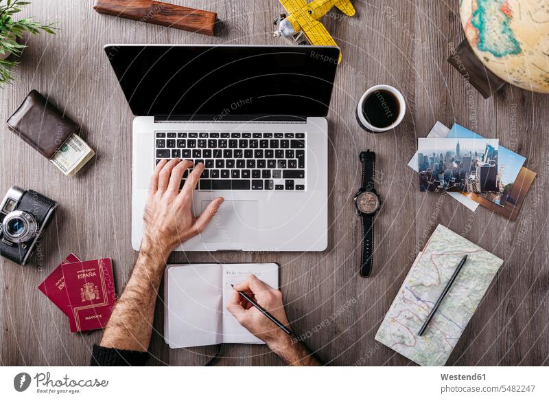 Draufsicht eines Mannes, der einen Laptop benutzt, umgeben von Reiseutensilien auf dem Tisch schreiben aufschreiben notieren schreibend Schrift Notebook Laptops