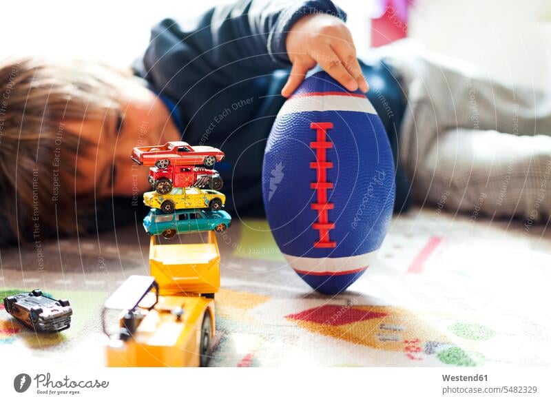 Auf dem Boden liegender Junge mit Fussball und einem Stapel Spielzeugautos Europäer Kaukasier Europäisch kaukasisch Faulheit faul Rugbyball Rugbybälle