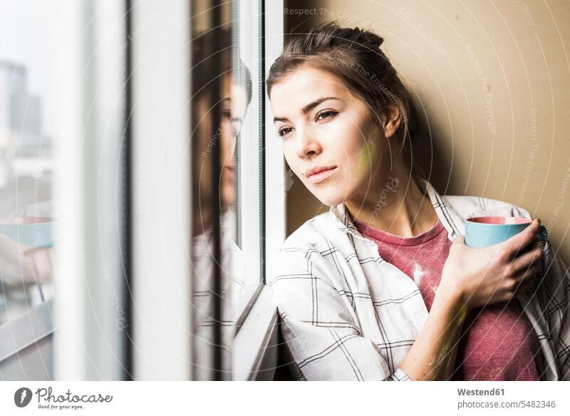 Junge Frau, die eine Kaffeetasse hält und aus dem Fenster schaut junge Frau junge Frauen Eigenheim Eigenheime wohnen renovieren Renovierung stehen stehend steht
