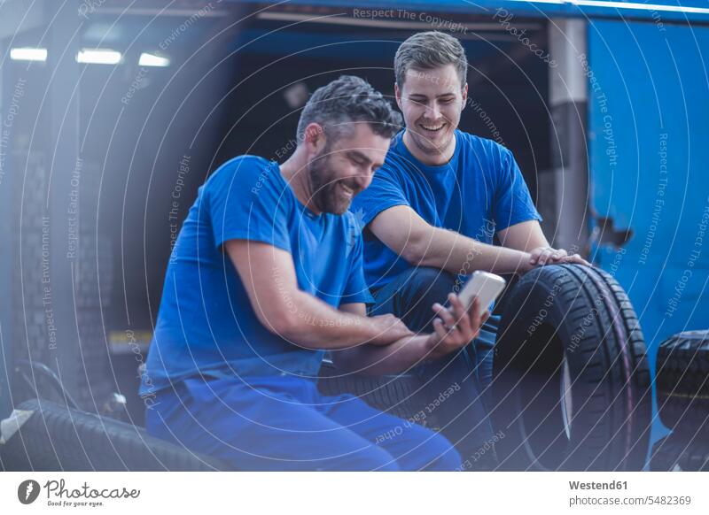 Zwei Mechaniker bei einer Pause mit Blick auf das Mobiltelefon lächeln sitzen sitzend sitzt Mann Männer männlich Reifen Handy Handies Handys Mobiltelefone