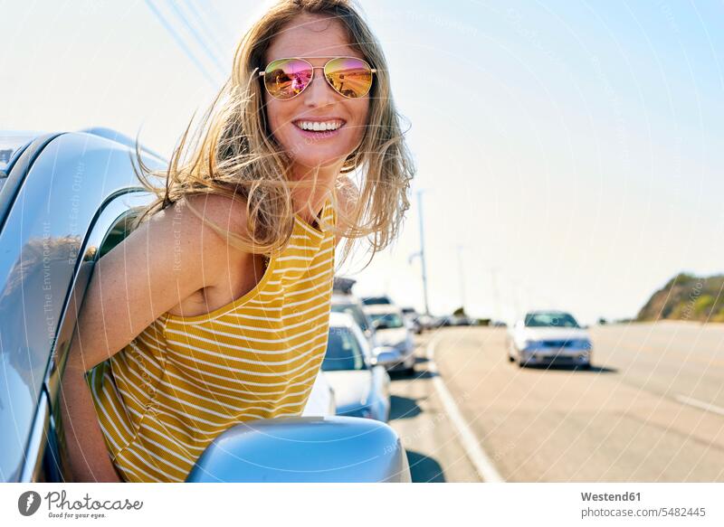 Glückliche junge Frau lehnt sich aus dem Autofenster Wagen PKWs Automobil Autos weiblich Frauen lächeln Kraftfahrzeug Verkehrsmittel KFZ Verkehrswesen