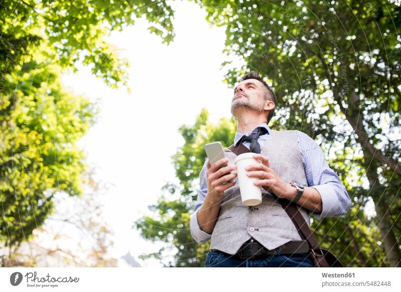Älterer Geschäftsmann mit Kaffee und Smartphone im Stadtpark Handy Mobiltelefon Handies Handys Mobiltelefone Mann Männer männlich Telefon telefonieren