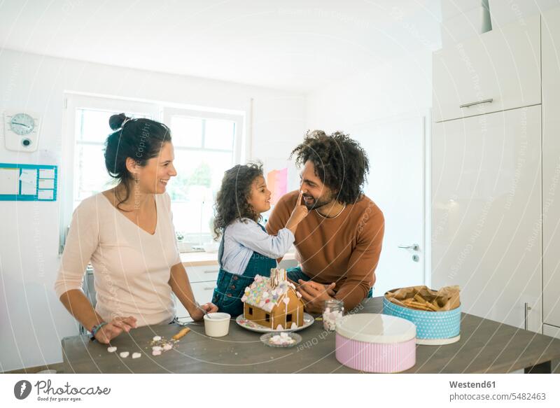 Familie backt zu Weihnachten Lebkuchenhaus mit Süßigkeiten, Tochter legt dem Vater Zuckerguss auf die Nase Lebkuchenhäuschen Lebkuchenhaeuschen Zuckerguß Nasen
