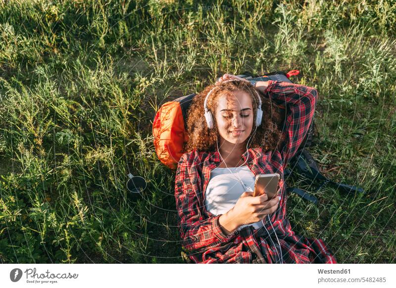 Teenagerin mit Rucksack, die auf einer Wiese mit Kopfhörern und Handy Musik hört liegen liegend liegt junges Mädchen Teenagerinnen weiblich junge Frau