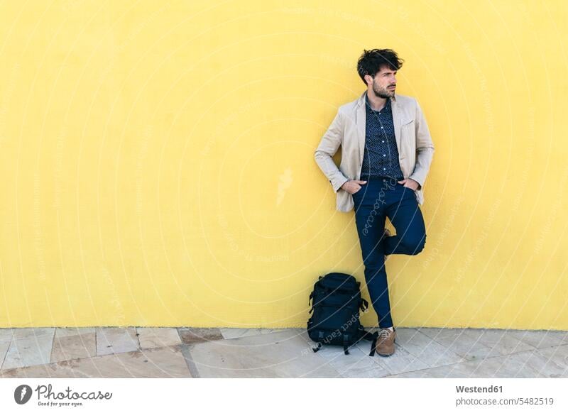 Junger Mann mit Rucksack vor gelber Wand stehend Männer männlich Erwachsener erwachsen Mensch Menschen Leute People Personen anlehnen angelehnt lehnend Wände