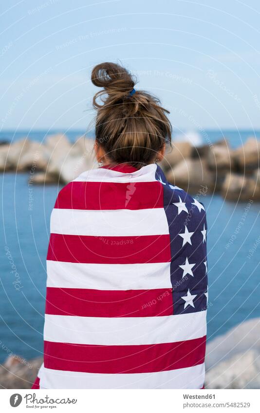 Rückenansicht einer jungen Frau, die in eine US-amerikanische Flagge gehüllt vor dem Meer steht weiblich Frauen Erwachsener erwachsen Mensch Menschen Leute
