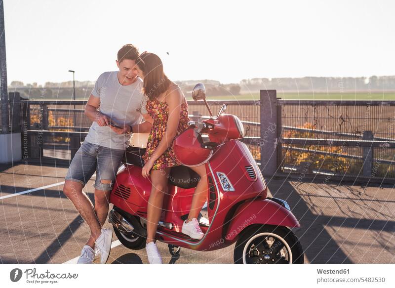 Glückliches junges Paar auf Parkebene mit Motorroller und Handy Mobiltelefon Handies Handys Mobiltelefone Parkdeck Parkdecks Pärchen Paare Partnerschaft