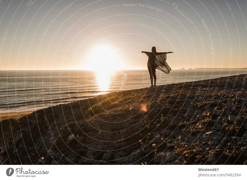 Rückenansicht eines glücklichen Teenager-Mädchens mit ausgestreckten Armen, das bei Sonnenuntergang auf einem Hügel steht und auf das Meer blickt Teenagerin